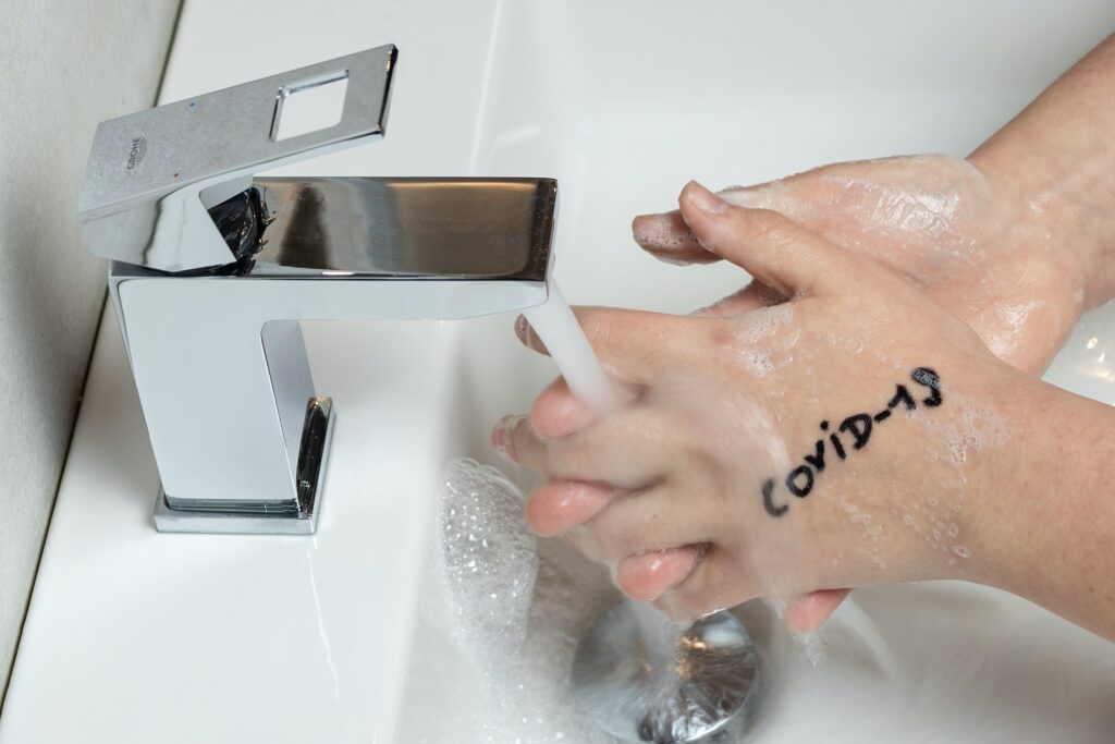 Hands being washed under the faucet; left hand facing camera has phrase COVID-19 written on the back of hand. Blog graphic for the blog post for awomansoutlook.com