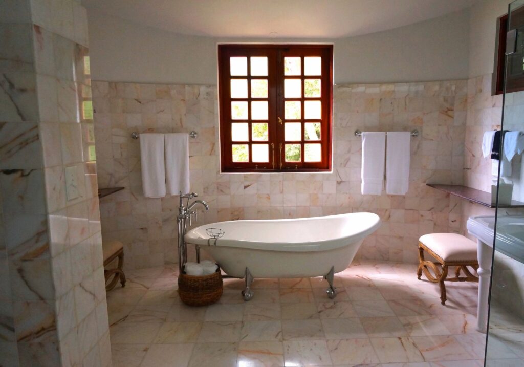Bathroom view with marble floor and most walls. Window with brown wooden trim. A towel rod with towels on either side of the window. Porcelain tub below window. Small bench is pushed under a table on side of tub next to tub. Glass shower stall to right of photo view. Blog graphic for blog post for awomansoutlook.com