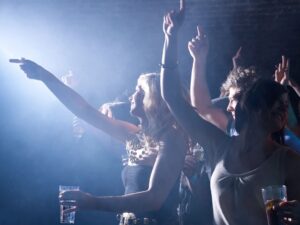 Photo of two females at a concert. One has her arms up over her head, cheering. The other is pointing to something outside of photo and has a cup filled with liquid in her other hand. Background is mostly dark, except there is a light shining toward them. Blog graphic for blog post How Alcohol Drinking Can Ruin A Night Out for awomansoutlook.com