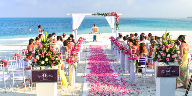 Beach side wedding with main aisle down the center with people in pews on either side of aisle. Minister and square wooden arch with white cloth draped. Red rose petals are sprinkled on white runner down to where minister is. Blog graphic for post three simple ways to enjoy a wedding ceremony from awomansoutlook.com