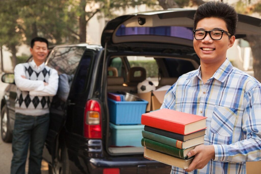Photo of young man with books in his hands standing next to the trunk of an SUV with boxed up items. Older man is standing beside the SUV. Blog graphic for blog post 10 true tips for every college freshman at awomansoutlook.com