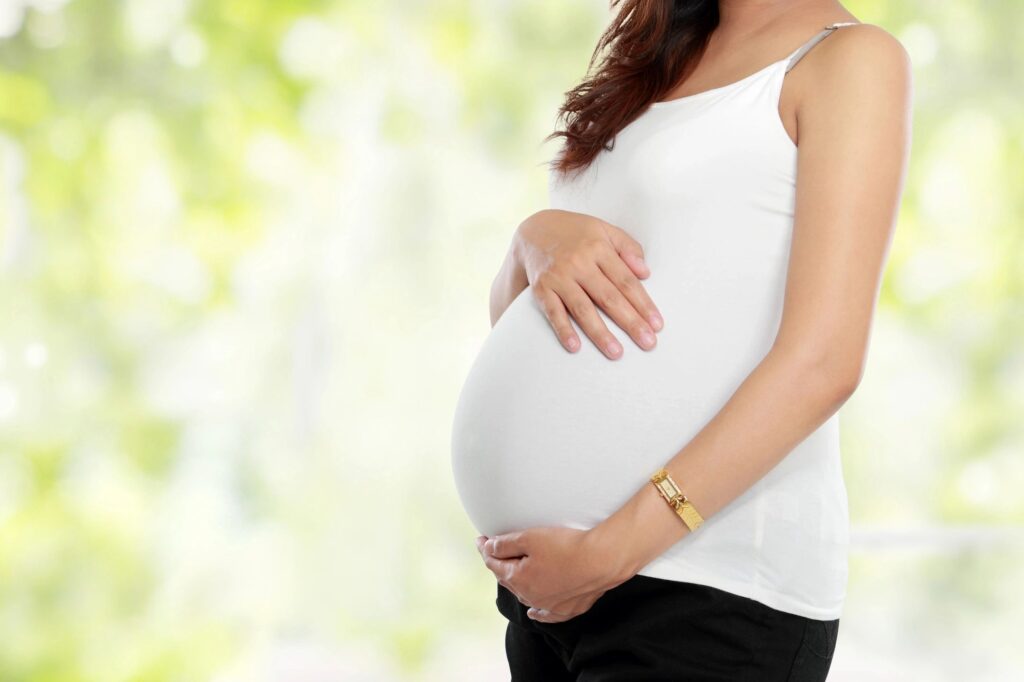 Photo of the side view of a pregnant woman's belly. The lady is wearing a white tank top. The background looks to be green trees, Blog graphic for blog post a simple guide on how babies are made at awomansoutlook.com